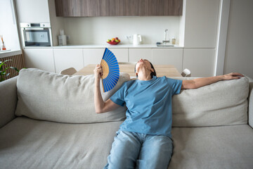Woman lounging on sofa fanning herself in hot airless room without air conditioning. Elderly discomfort from sweltering, heatwave vulnerability, poor ventilation struggle, aging female hardship