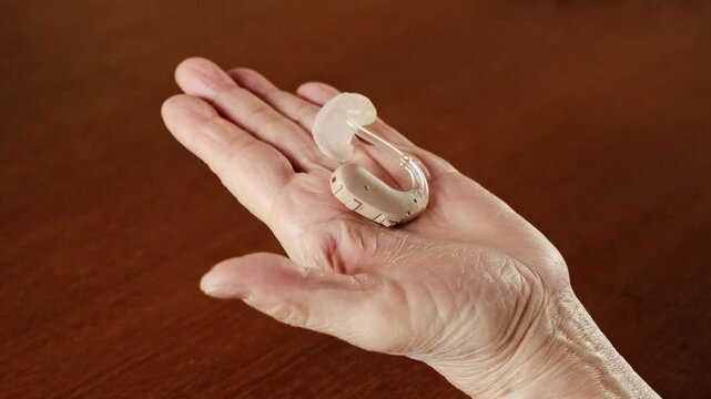 Close-up of a senior hand gently holding a modern hearing aid device over a wooden table, illustrating hearing assistance technology, elder care tools, and accessibility in healthcare solutions.