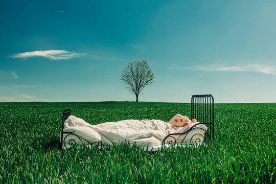 Woman dreaming in a bed outdoors under a blue sky with a lonely tree in a green field
