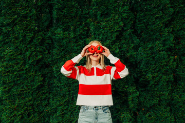 Woman in a striped shirt holding a tomato and bell pepper outdoors in front of arborvitae