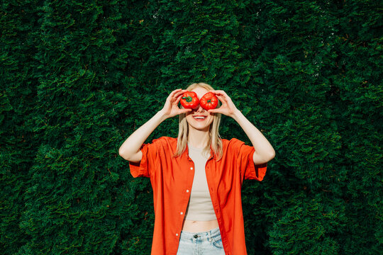 Woman in a red shirt holding a tomato and bell pepper in front of her eyes outdoors with a green arborvitae background