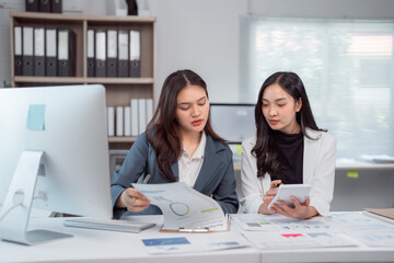 Two asian businesswomen are working together at modern office desk. Analyzing financial charts. Discussing and using calculator. Collaborating on investment strategy and planning for corporate growth