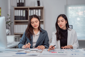 Two young businesswomen collaborating in a modern office, analyzing financial charts and reports on...