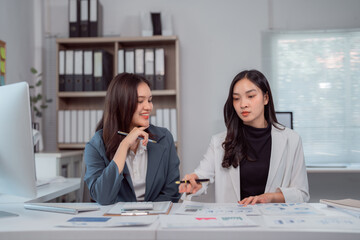 Two young businesswomen are collaborating on a project, analyzing financial charts and discussing...