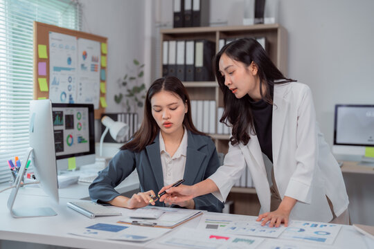 Two Asian businesswomen discussing and analyzing financial data and charts while using a calculator and computer in a modern office setting, exemplifying teamwork and collaboration