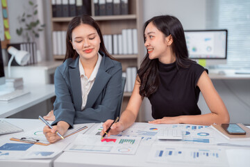 Two Asian businesswomen collaborating at a modern office desk, analyzing financial charts and...