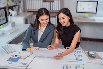 Two young Asian businesswomen are examining financial documents and discussing data analysis in a...