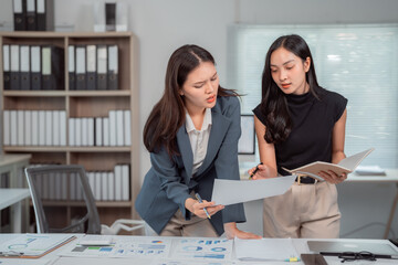 Two asian businesswomen reviewing financial reports and pointing at charts and graphs while discussing company performance and making strategic decisions in modern office