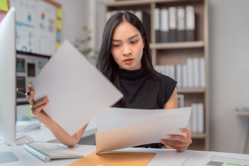 Fototapeta premium Young Asian businesswoman reviewing documents and working on a computer at an office desk, focused on analyzing financial data and checking reports