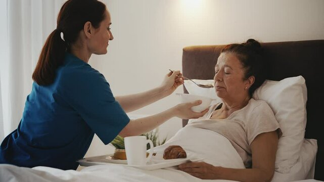 Nurse spoon-feeding elderly woman lying in bed, assisted living, home care