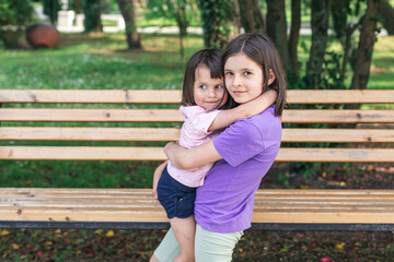 Two sisters share a warm hug in a park. The older girl holds the younger one in her arms as they both look at the camera, standing in front of a wooden bench surrounded by greenery.