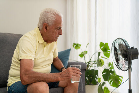 Senior man suffering from summer heatwave holds glass of water while sitting on the sofa near a fan