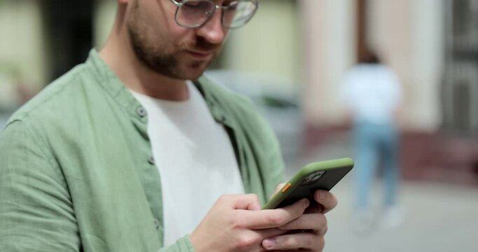 Portrait of casual man in glasses using smartphone on the street. Handsom bearded man typing messages on phone outside. Online, messaging and browsing internet.