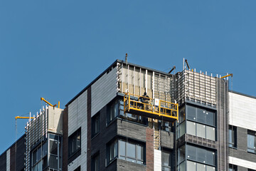 Construction worker on suspended platform repairs high-rise building facade against clear blue sky, modern urban development scene. © SOLDATOOFF