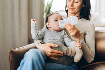 Loving mother feeding baby with milk bottle on sofa
