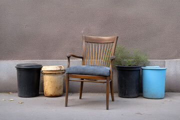 Naklejka premium Vintage wooden chair with a blue cushion is positioned between various trash bins against a textured wall, creating an intriguing urban scene with contrasting elements