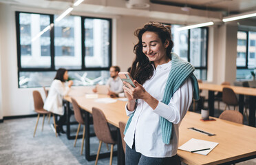 Smiling young woman checking smartphone with joy during office break, relaxed and casual energy in modern workspace, colleagues blurred in background, warm daylight