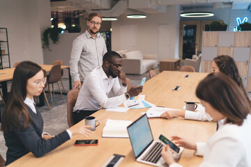 Focused group of coworkers at long desk using smartphones and laptops, engaged in multitasking...