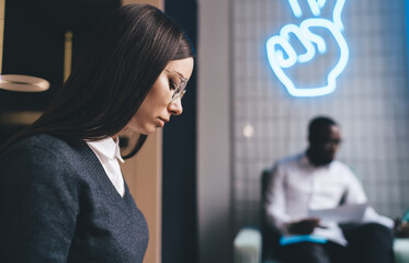Focused young businesswoman reading document in stylish office with neon sign and colleague in...