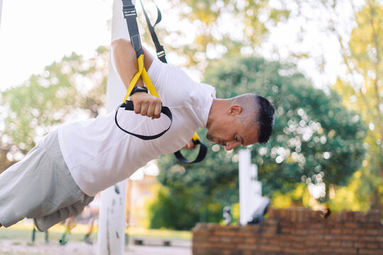 Muscular athletic powerful man bodybuilder sportsman use sport equipment outdoor stretching workout on city sports ground hanging pull ups exercise with TRX straps active healthy life