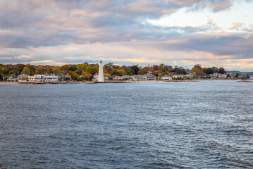 Lighthouse and waterfront residential properties along a rugged coast bordered by colourful autumn trees in Connceticut  under cloudy sky at sunset