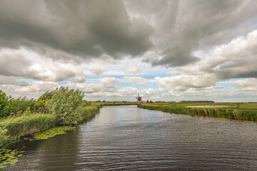 A characteristic Dutch polder landscape featuring a historic thatched hollow post windmill, set beside a wide canal. Captured on a windy spring day under a dramatic sky filled with dark clouds