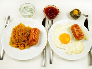 Plates with grilled sausages, stewed cabbage and fried eggs as a garnish are on a white table. Table setting, lunch, dinner