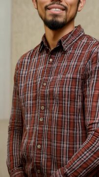 Smiling young adult with glasses and beard wearing a button-down flannel shirt indoors against a neutral background in a close-up portrait