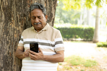 Fototapeta premium Indian Active happy senior man using and text messaging on mobile while standing at park. Wearing red sweet sweater and eyewear sun glasses. Indian Senior man using digital technology at outdoors