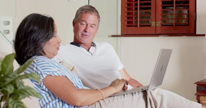 Senior diverse couple relaxing on sofa, smiling while using laptop at home