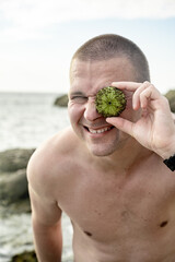 A cheerful funny guy holds a sea urchin shell near one eye. Summer fun and entertainment concept