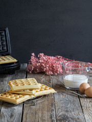 freshly made homemade waffles on a rack, ingredients on the side and waffle iron on the bottom. Rustic wooden table in a Nordic and intimate atmosphere.