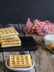 Stack of freshly made waffles on a rustic wooden table, with ingredients and waffle maker in the background. Cozy homemade breakfast scene in natural light