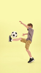 Boy lifting leg to balance ball in classic soccer move, wearing shorts and sneakers, yellow background enhances focus on movement and coordination. Concept of summer activities, hobby, holidays
