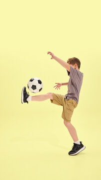 Fototapeta Boy lifting leg to balance ball in classic soccer move, wearing shorts and sneakers, yellow background enhances focus on movement and coordination. Concept of summer activities, hobby, holidays