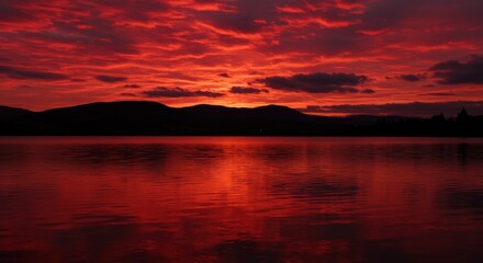 Naklejka premium Fiery sunset reflecting on a tranquil lake with mountain silhouettes