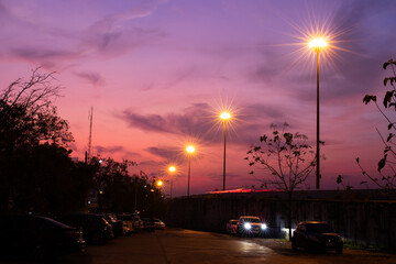 Streetlights glow under a dramatic sunset sky with purple and pink tones. Silhouettes of trees, cars, and people create a calm urban evening atmosphere perfect for background and editorial use.