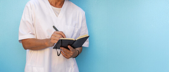 A nurse in a white uniform takes notes in a black notebook with a pen, against a uniform blue background. The image is ideal for banners or headers. Copy Space.