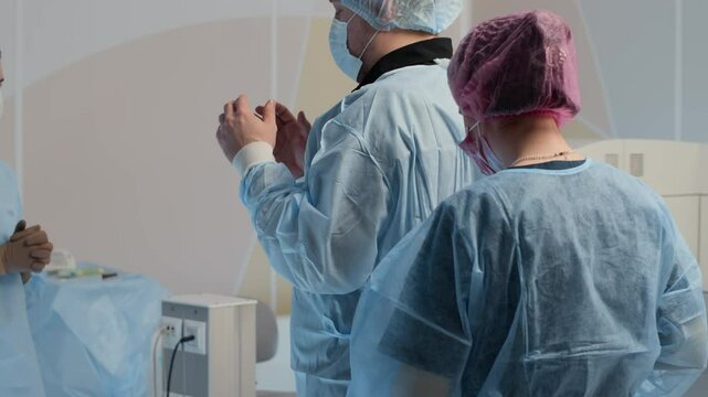 Medical staff dressed in full protective attire, including gowns, gloves, and masks, preparing surgeon for procedure in sterile operating room