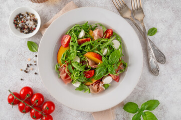 Salad with peaches, arugula, prosciutto, mozzarella cheese and cherry tomatoes with balsamic vinegar in a ceramic plate on a concrete background. Selective focus.