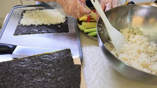 Making a Kimbab, Korean rice roll. Work station with cutting board, sheets of dried seaweed, bowl of rice and a plate full of kimbab fillings on a table