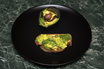 Avocado halves and avocado toast with mashed avocado on a dark plate background. Healthy food and simplicity in modern culinary aesthetics.