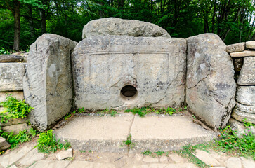 Stone dolmen with circular opening, surrounded by lush forest. Large rocks form ancient structure,...
