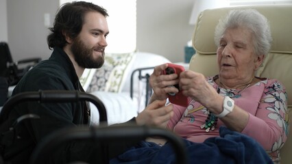 Obraz premium A young man joyfully shows his grandmother pictures on a mobile device as they share treasured memories, creating a moment of happiness together.