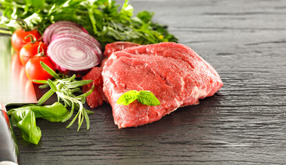 Raw seasoned pieces of raw meat prepared for cooking, placed on a kitchen table surrounded by fresh ingredients and herbs  – healthy cooking and lifestyle concept. 