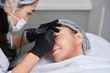 Skilled technician applies permanent eyebrow makeup to young woman in calm salon. With black...