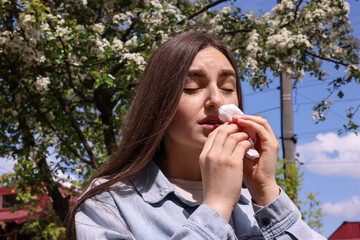 Woman with paper tissue suffering from seasonal pollen allergy near blossoming tree on spring day