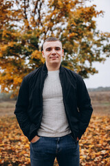 Smiling young man standing outdoors in an autumn park. He is wearing a black jacket and jeans, posing confidently in front of a tree with vibrant orange and yellow leaves. Casual and cheerful mood.