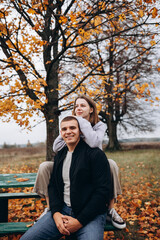 Fototapeta premium Young couple sitting and hugging on a bench under a yellow-leaved tree. Autumn idyll in nature, a romantic moment among fallen leaves in a field. A warm, cozy atmosphere surrounded by colorful leaves 