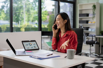 Fototapeta premium Asian businesswoman talking on mobile phone while working on laptop at office desk. communication, and corporate success.